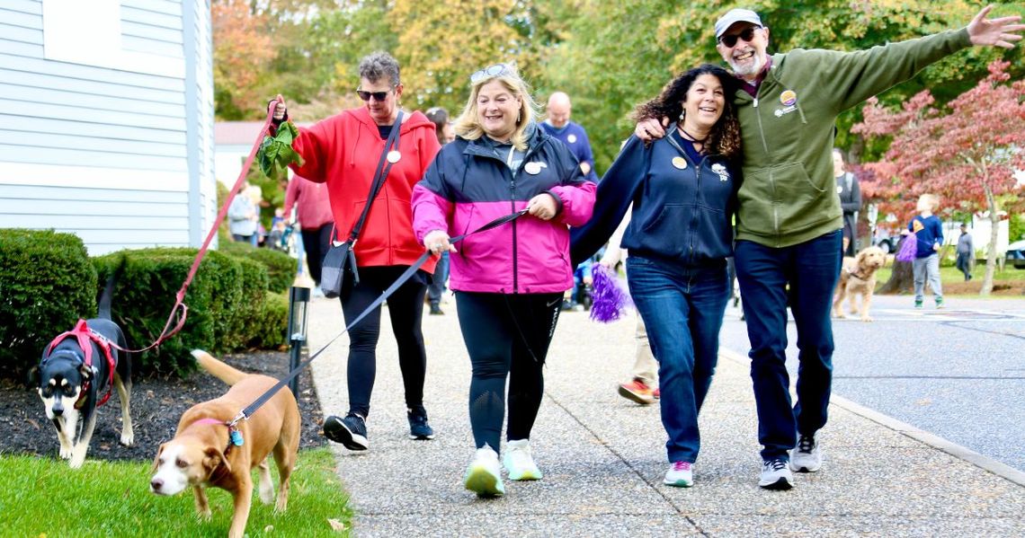 A group of people and their dogs walk the 3 mile Interfaith Mini Walk for Hunger route in Westwood, MA and smile for the camera.