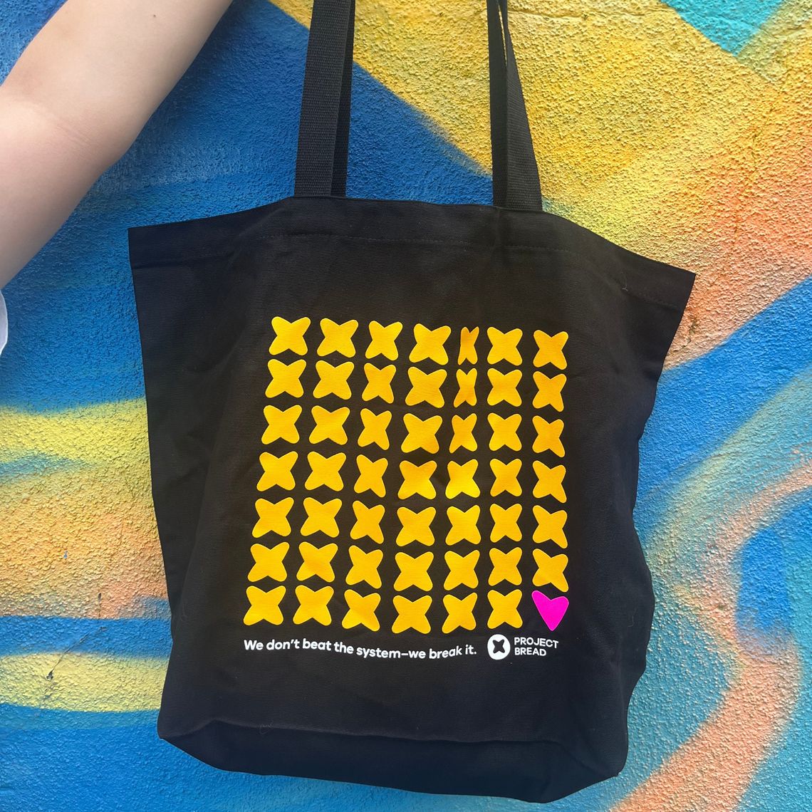 Black tote bag with project bread x's and a heart that says "We dont beat the system we break it" A sticker set with two stickers, one in the shape of bread that says"Champion of Change, I give monthly to Project Bread" the second is a yellow heart that says "Monthly DONOR committed to ending hunger" with a project bread logo