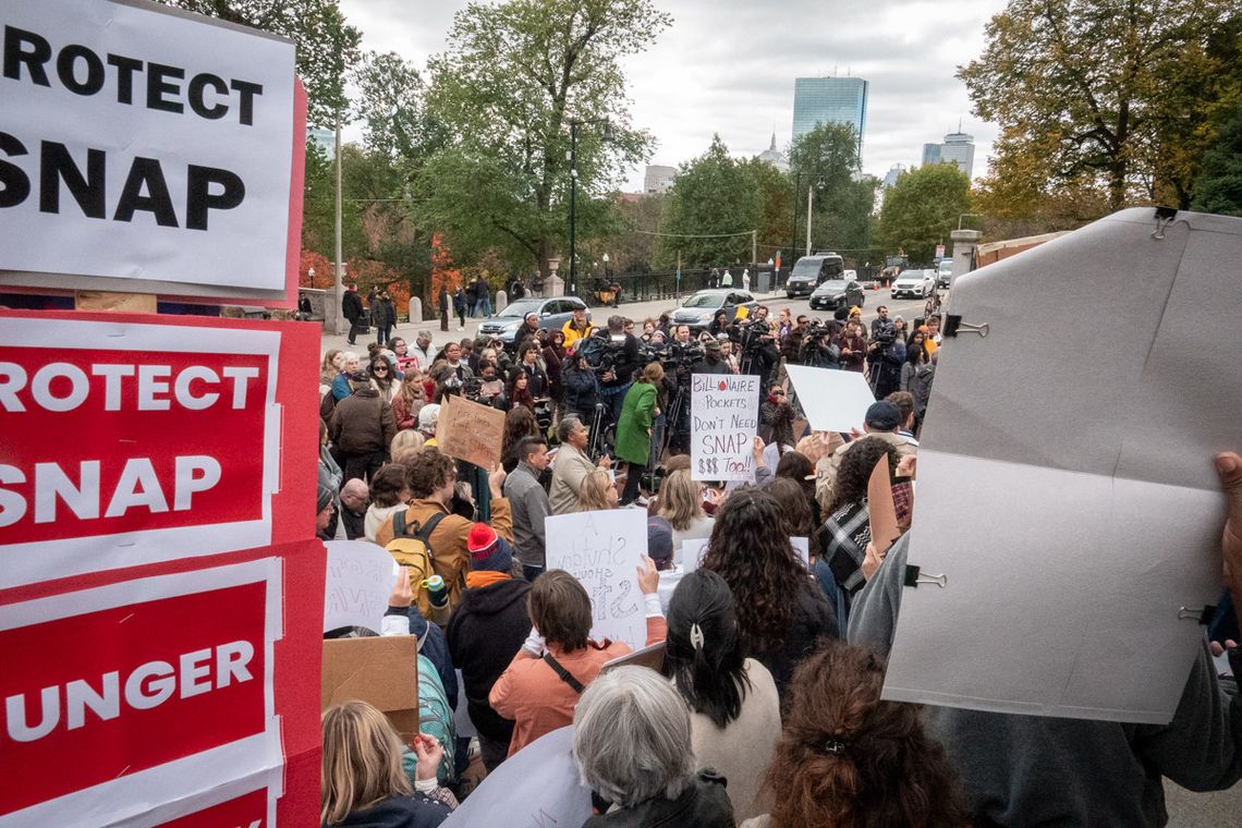 A large crowd holds signs that say "Protect SNAP" and listens to speaker at the State House Steps