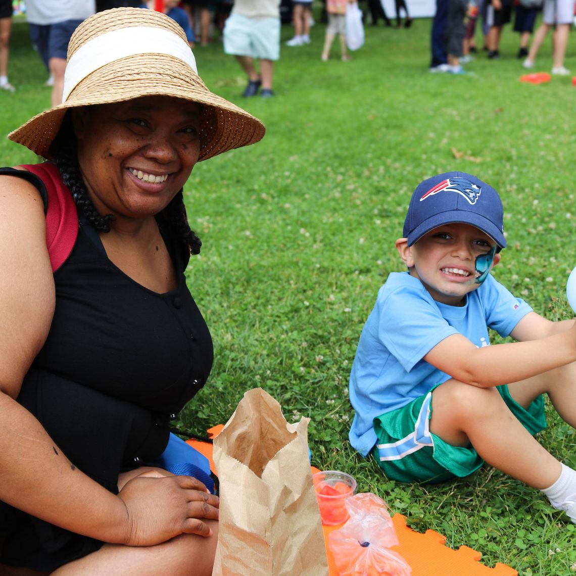 A Waltham parent and her child sit down in the shade at the Summer Eats Kickoff in Waltham.